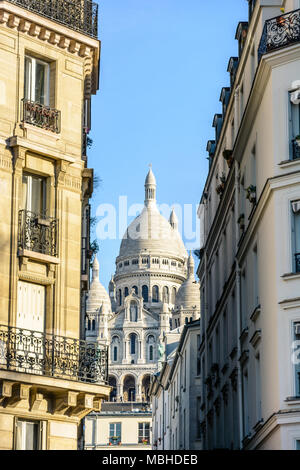 Blick auf die Fassade und die Kuppel der Basilika des Heiligen Herzens in Paris durch eine enge Straße zwischen typischen Gebäude unter einem klaren blauen Himmel. Stockfoto