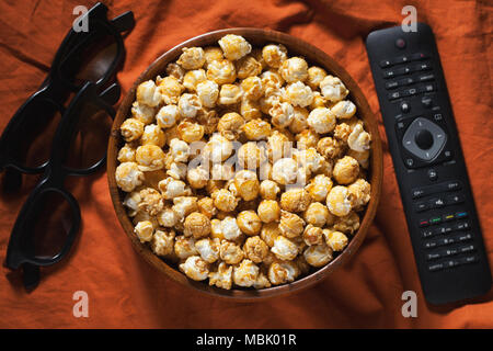 Hölzerne Schüssel mit süßem Popcorn, TV-Fernbedienung und 3D-Brille auf orange Bettwäsche. Ansicht von oben. Snacks und Essen für einen Film Stockfoto