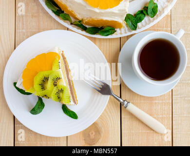 Ein Stück Obst Kuchen (Kiwi, Orange, Minze) auf einer weißen Platte auf Holz- Hintergrund und einer Tasse Tee. Die Ansicht von oben Stockfoto