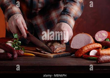 Mann in einem brown Plaid Shirt schneidet verschiedene Wurst und geräuchertes Fleisch auf einem alten Holztisch. Stockfoto