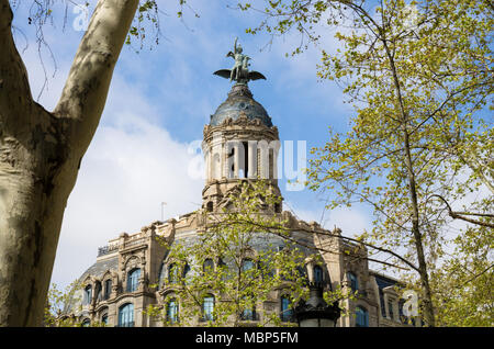 Bäume rahmen den Blick auf eine Abbildung mit einem geflügelten Vögel sitzen auf der Spitze der Kuppel an der Spitze eines Gebäudes auf dem Passeig de Gràcia in Barcelona, Spanien. Stockfoto