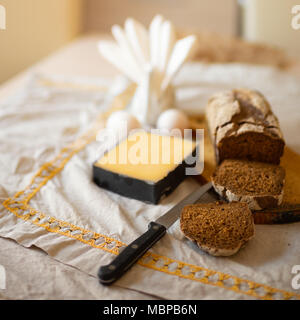 Ein Laib Brot mit in Scheiben geschnittenen Stücke auf einem Schneidebrett mit Messer, eine Scheibe Käse und Eier liegen auf einem Tisch mit einem Bettwäsche licht Tischdecke Stockfoto