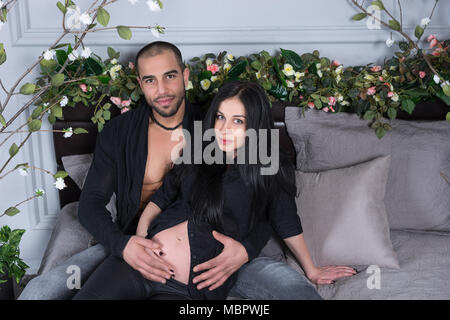 Stattliche muslimischen Mann und seine schwangere Frau tragen schwarze Kleidung, er umarmt ihren Bauch im Sitzen auf der grauen gemütliches Bett im Schlafzimmer Stockfoto
