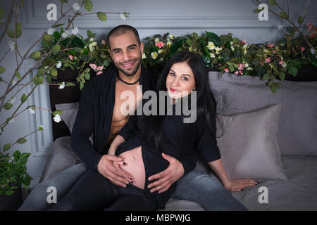 Wunderbare paar muslimischen Mann und seine schöne schwangere Frau tragen schwarze Kleidung, er umarmt ihren Bauch und lächelte, während er auf den grauen gemütliche Stockfoto