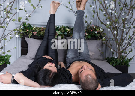 Wunderbare paar muslimischen Mann und seine schwangere Frau tragen schwarze Kleidung, hob ihre Beine auf, während er sich auf den grauen gemütliches Bett im Schlafzimmer liegen Stockfoto