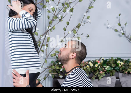 Friedliche internationale Paar in gestreiften Pullover sehen einander an, muslimischen Mann setzte sich und umarmt seine schwangere Frau im Schlafzimmer Stockfoto