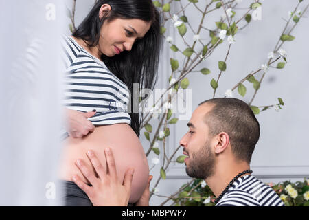 Schöne internationale Paar in gestreiften Pullover sehen einander an, muslimischen Mann setzte sich gegenüber und dem Bauch seiner schwangeren Frau Holding in der Stockfoto
