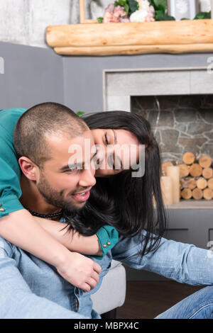 Gerne internationale Paar Mann mit Bart und seinen brunette schwangere Frau auf dem Sofa sitzen und ihn umarmen und küssen in die Wohnzimmer mit einem Stockfoto