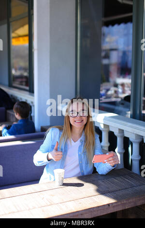 Mädchen zeigen Daumen oben, sitzen an Street Cafe mit Smartphone. Stockfoto