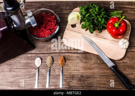 Elektrischer Fleischwolf in der Nähe von eine Platte mit Hackfleisch, Knoblauch, Zwiebeln, Paprika, Pfeffer, Petersilie, Messer und Löffel mit Gewürzen auf einer hölzernen Hintergrund, Konz Stockfoto