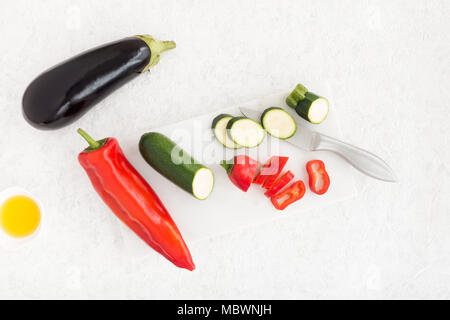 Aubergine, Paprika und Zucchini süß wies auf weißem Marmor Schneidebrett mit einer Schüssel mit Olivenöl auf Weiß marmorierten Hintergrund. Ansicht von oben. Stockfoto