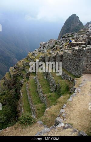 Mann sitzt auf der Kante einer Wand an der verlassenen Zitadelle von Machu Picchu hoch in den peruanischen Anden und hinunter ins Tal Stockfoto