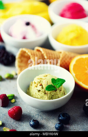 Eis Kugeln in Schalen, Waffeln, Beeren, Orange, Mango, Pistazien auf grauem Beton Hintergrund. Bunte Sammlung, Flach, Sommer Konzept Stockfoto