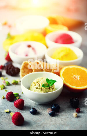 Eis Kugeln in Schalen, Waffeln, Beeren, Orange, Mango, Pistazien auf grauem Beton Hintergrund. Bunte Sammlung, Flach, Sommer Konzept Stockfoto