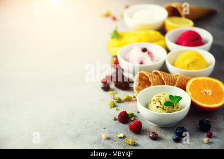 Eis Kugeln in Schalen, Waffeln, Beeren, Orange, Mango, Pistazien auf grauem Beton Hintergrund. Bunte Sammlung, Flach, Sommer Konzept Stockfoto