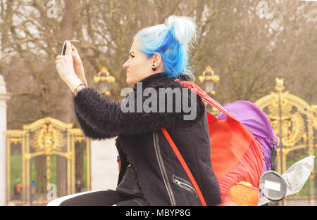 Eine junge Frau mit markanten blauen Haare ein Foto auf Ihrem Smartphone mit zwei bunten Rucksäcke vor dem Buckingham Palace Stockfoto