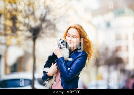 Junge rothaarige kaukasische Frau mit Sommersprossen im Gesicht hält und Küsse, Umarmungen, liebt Schwarze und Weiße shaggy Hund Chihuahua Zucht. Das Mädchen gekleidet in Stockfoto