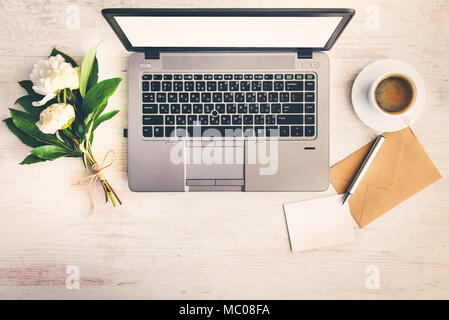 Blick von oben auf einen Schreibtisch mit Computer, Kaffeetasse, Kraftpapier Umschlag, leerer Hinweis und ein Bündel von Pfingstrose Blüten mit weißem Holz Hintergrund. Stockfoto