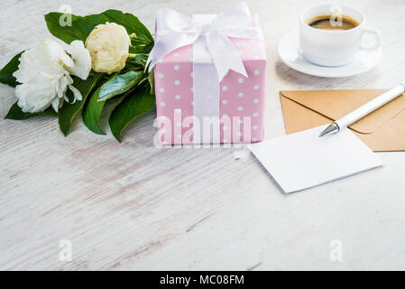Pink gepunktete Geschenkbox, leere Grußkarte, Kraftpapier Umschlag, Pfingstrosen Bouquet und Kaffee Tasse über weißen Holzmöbeln rustikal Tisch. Romantischen Urlaub und Fall c Stockfoto