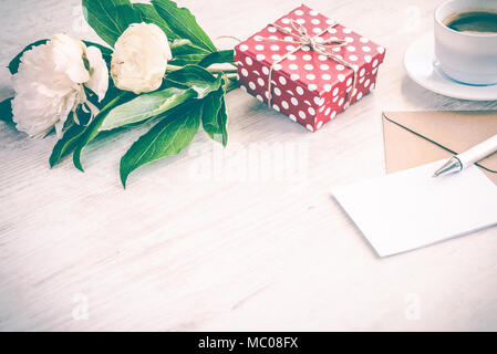 Gepunktete rote Geschenkbox, Kraftpapier Umschlag und leere Grußkarte, Kaffeetasse und Pfingstrose blumen blumenstrauss über weiß Holz Hintergrund. Kopieren Sie Platz. Vintage Effe Stockfoto