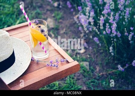 Ein Glas erfrischende Limonade und einen Strohhut über einen hölzernen Stuhl. Blühende Lavendel Blumen im Hintergrund. Stockfoto