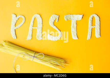 Das Wort "Pasta wird in Massen Briefe von kleinen rohen Makkaroni auf gelbem Hintergrund legte neben einem Haufen trockene Spaghetti. Essen auf einem hellen Hintergrund Stockfoto