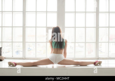 Junge schöne Mädchen mit einem blauen Hemd und weißen Shorts führt Gymnastik element Garn. Sie sitzt auf einer Fensterbank, dahinter befindet sich ein Fenster. Stockfoto