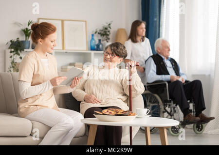 Krankenschwester, ein Buch lesen, eine ältere Frau mit einem anderen Rentner sitzt in einem Rollstuhl im Hintergrund Stockfoto
