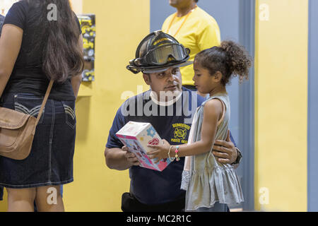 HUMACAO, Puerto Rico, 6. Januar 2018 - Feuerwehrmann Hände Geschenk zu einem kleinen Mädchen, das die Großen Drei König Feier am Humacao Arenas Kolosseum besucht. Verwaltung der psychischen Gesundheit und Suchtprävention (ASSMCA), das in Zusammenarbeit mit dem Außenministerium, FEMA, und anderen Einrichtungen nahmen an der großen Feier der Heiligen Drei Könige in Aguada, Orocovis, Humacao, Manati, Salinas, und Guaynabo, zum Nutzen der Freude und Geschenke an Kinder und ihre Familien. ASSMCA soll helfen, die emotionale Belastung, die der Hurrikan Maria unter Beibehaltung der tradit verringern Stockfoto