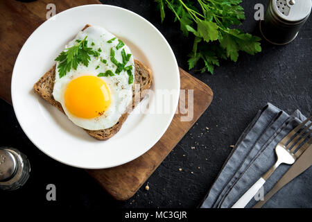 Spiegelei auf Toast zum Frühstück. Spiegelei mit Brot und Petersilie auf weiße Platte auf Schwarz, Ansicht von oben, kopieren. Stockfoto