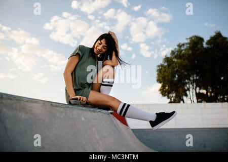 Weiblichen Skateboarder, eine gute Zeit bei Skate Park. Gerne Frau sitzt auf Skateboard Rampe im Freien und lächelnd. Stockfoto