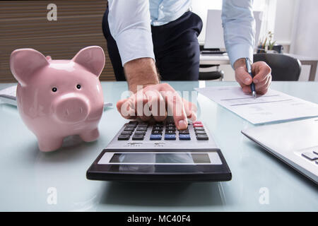 Unternehmer mit Taschenrechner für die Berechnung der Rechnung mit Sparschwein auf dem Schreibtisch Stockfoto