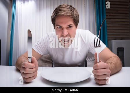 Wütend Mann mit Messer und Gabel mit leeren Teller am Tisch Stockfoto