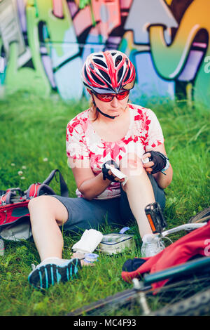 Frau Abrichten der Wunde auf ihr Knie mit Medizin in Spray und Gaze auf Radtour im Sommer Tag Stockfoto