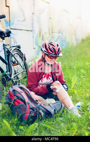 Frau Abrichten der Wunde auf ihr Knie mit Medizin in Spray und Gaze auf Radtour im Sommer Tag Stockfoto