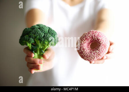 Junge Frau im weißen T-Shirt Wahl zwischen Brokkoli oder Junk Food, Donut. Gesunde, saubere detox Essen Konzept. Vegetarisch, vegan, raw-Konzept. Platz kopieren Stockfoto