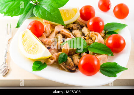 Salat von blanchierte Stücke von Meeresfrüchten auf einer weißen Platte close-up. Muscheln und Tintenfisch, dekoriert mit grünen, Cherry Tomaten und Zitronen mit Stockfoto