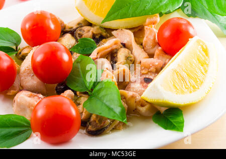 Salat von blanchierte Stücke von Meeresfrüchten auf einer weißen Platte close-up. Muscheln und Tintenfisch, dekoriert mit grünen, Cherry Tomaten und Zitronen mit Stockfoto