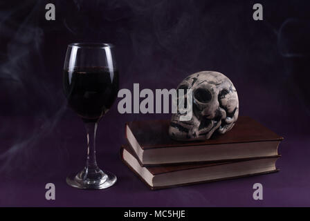 Schädel auf Bücher und Wein Glas auf lila dunkel und geräuchertem Hintergrund, Halloween Konzept, Low Key Stockfoto