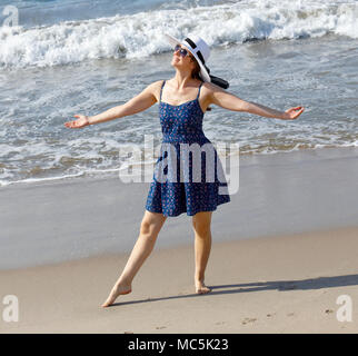 Junge Frau am Strand in einer freudigen Pose mit Arme weit offen. Er trägt einen blauen sundress, eine weiße Sonnenhut und Sonnenbrille Stockfoto