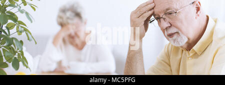 Nahaufnahme einer besorgt, älterer Mann tief in Gedanken versunken, mit der Hand auf seine Stirn und eine traurige ältere Frau unscharf im Hintergrund Stockfoto