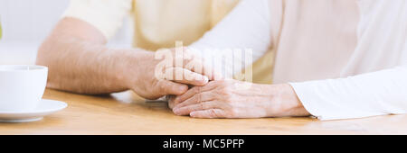 Eine Nahaufnahme der faltigen Armen auf einem Tisch, ein älterer Mann seine Hand auf den Händen eine ältere Frau Zuneigung zeigen und ihre Unterstützung der Holding Stockfoto