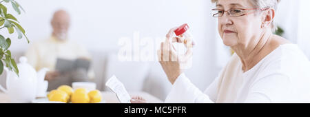 Eine ältere Frau Brille auf eine durchsichtige Flasche mit Pillen suchen und halten eine Droge Beipackzettel. Eine unscharfe Abbildung eines älteren Mannes lesen Stockfoto