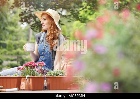 Hübsche Frau mit Strohhut auf dem Kopf Tee trinken auf der Terrasse sitzend neben Containern mit roten Blumen Stockfoto