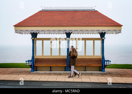 Zwei Menschen laufen entlang der Oberseite der Whitby West Cliff Bereich hinter einer Sitz Regenschutz am 12. April 2018 Stockfoto