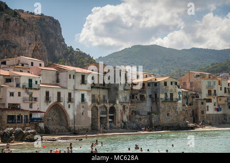 Felsigen Landzunge und historischen Gebäude mit Blick auf den Sandstrand von Cefalu, Sizilien, Italien, mit Urlaubern im Meer schwimmen. Stockfoto
