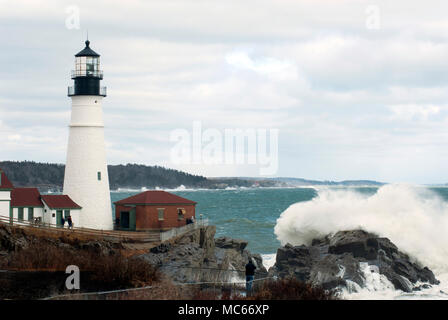 Große Wellen brechen über schroffe Felsen durch Portland Head Lighthouse in Maine an einem bewölkten Tag im späten Winter Saison. Touristen schauen vorwärts. Stockfoto