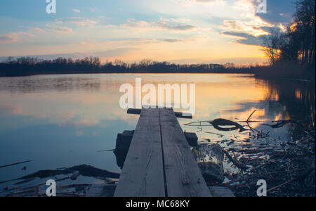 Sprungbrett am Ufer des Sees in der untergehenden Sonne. Schöne idyllische Szene - Sonnenuntergang über dem See. Wolken und Bäume spiegeln sich auf der Wasseroberfläche. Ar Stockfoto