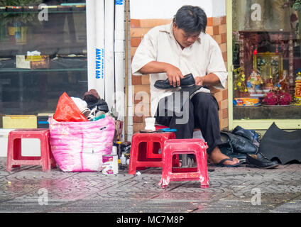 Ein Mann sitzt auf einem kunststoffhocker Schuhe reparieren, arbeiten auf dem Gehweg auf einer Straße in Ho Chi Minh City, Vietnam. Stockfoto