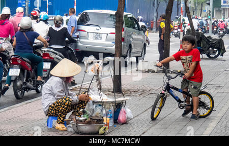 Eine Frau trägt einen konischen hat kochen Waffeln auf dem Gehweg auf einer belebten Straße in Ho Chi Minh City, Vietnam. Stockfoto